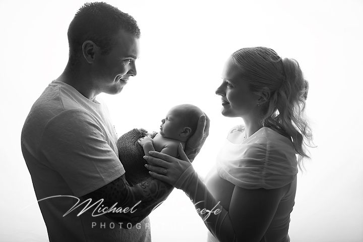 Black and white photo of parents lovingly holding their newborn baby, with soft backlit glow, by Michael Korfe Photography.