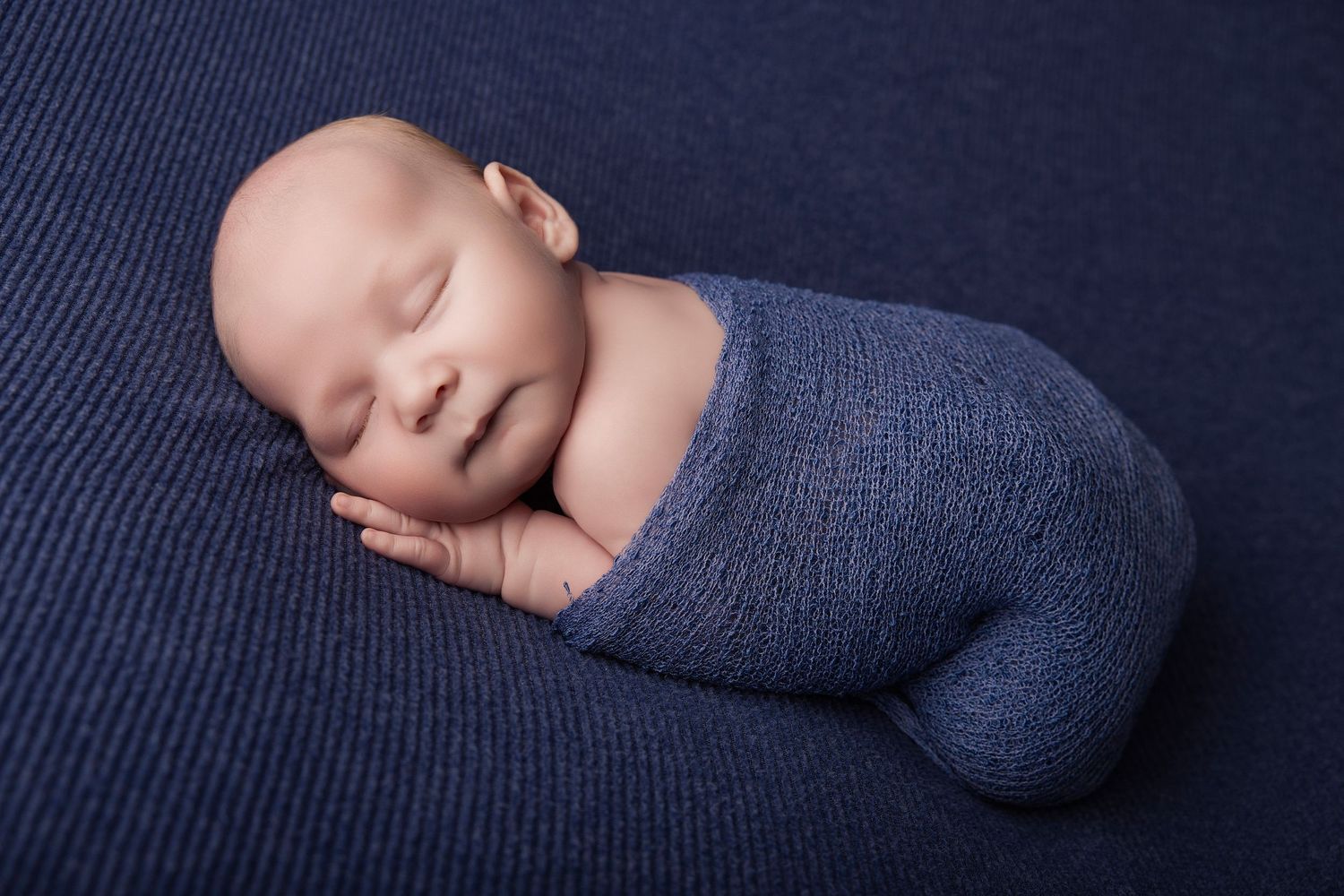 Peaceful newborn baby swaddled in a blue knit wrap, sleeping on a navy textured blanket.
