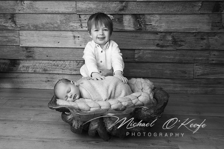 Black and white photo of toddler boy smiling behind sleeping newborn in wooden bowl by Michael O'Keefe Photography.
