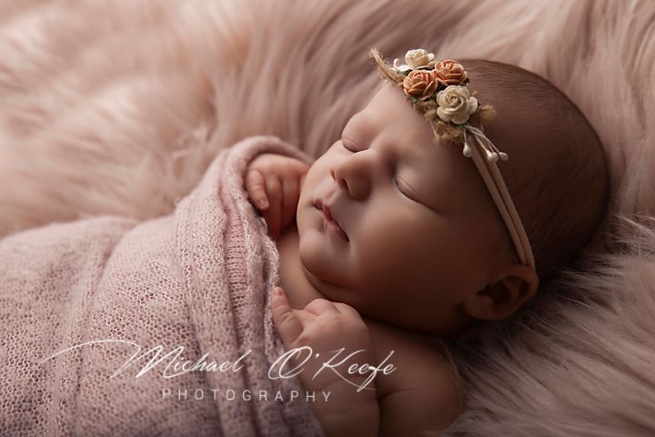Sleeping newborn baby wrapped in pink with a floral headband on a soft pink fur backdrop.
