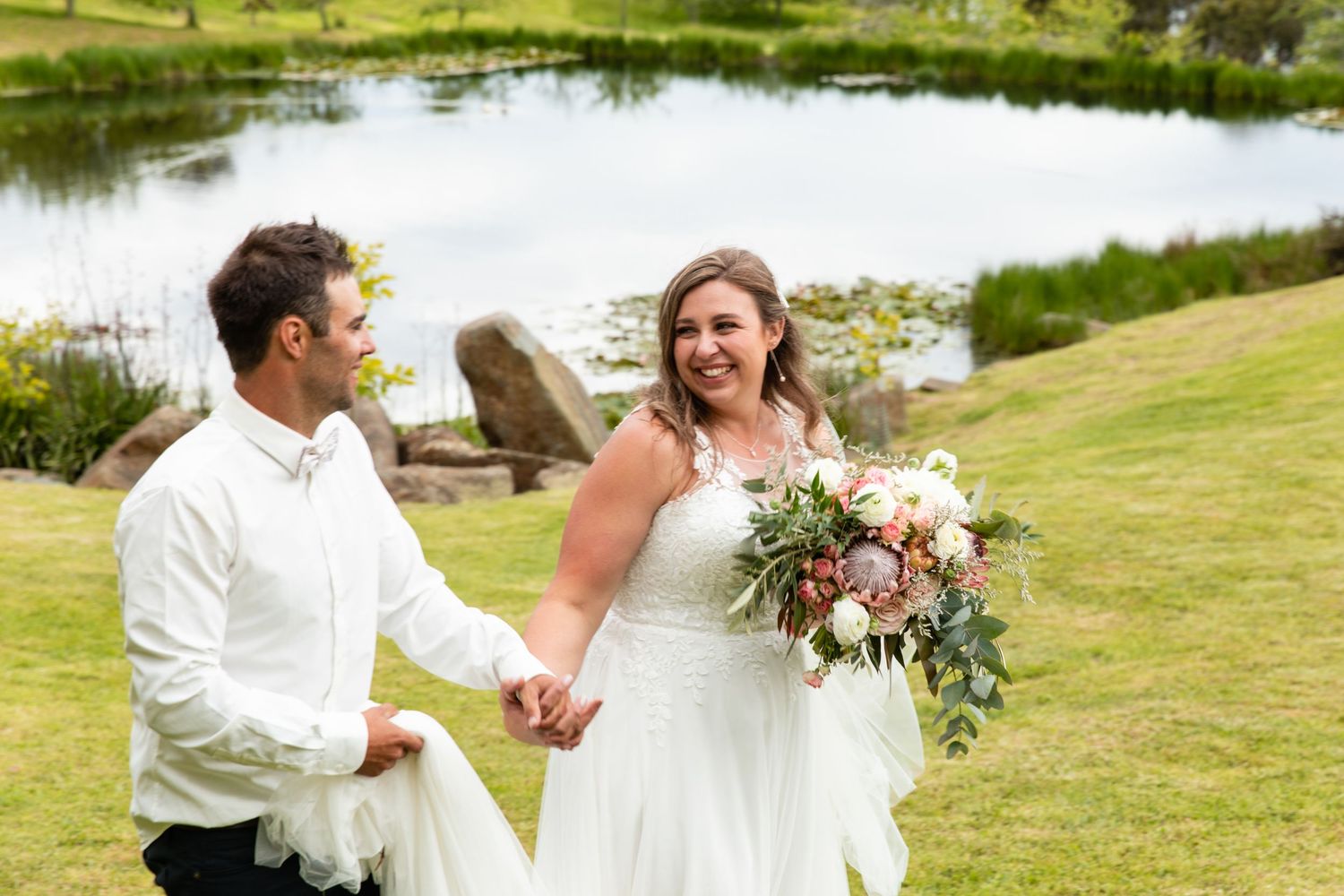 A couple shares a joyful moment by a pond with the bride holding a beautiful floral bouquet.