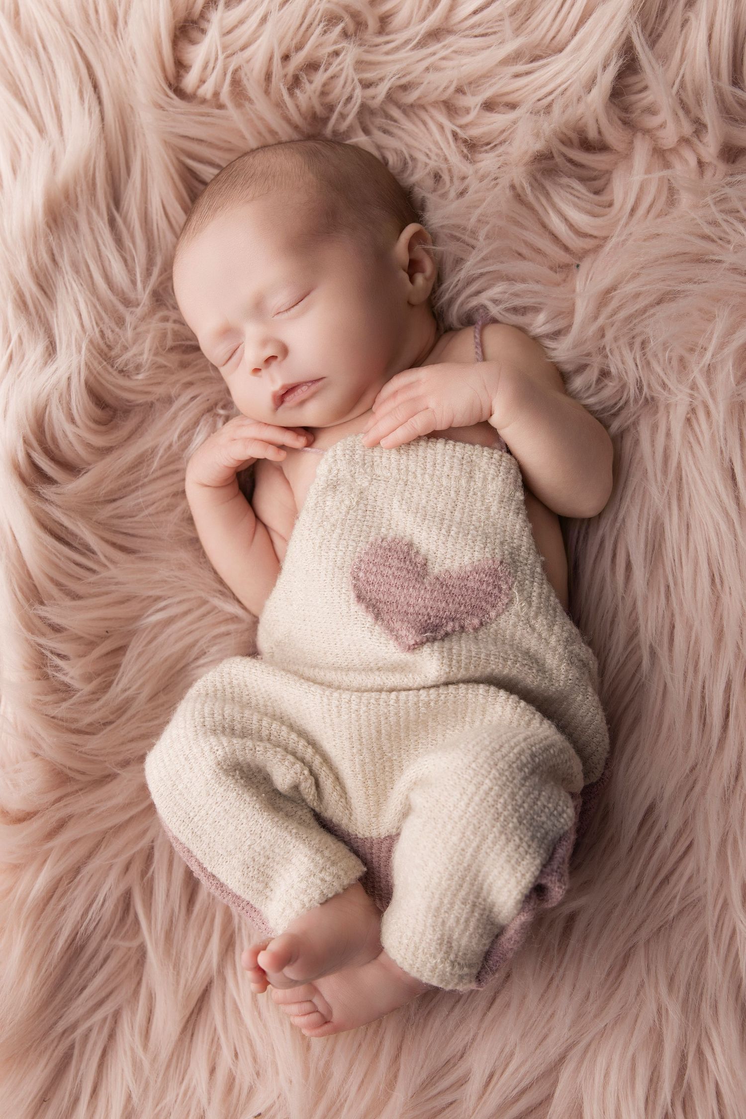 Sleeping newborn in knit romper with heart detail on pink fluffy blanket.