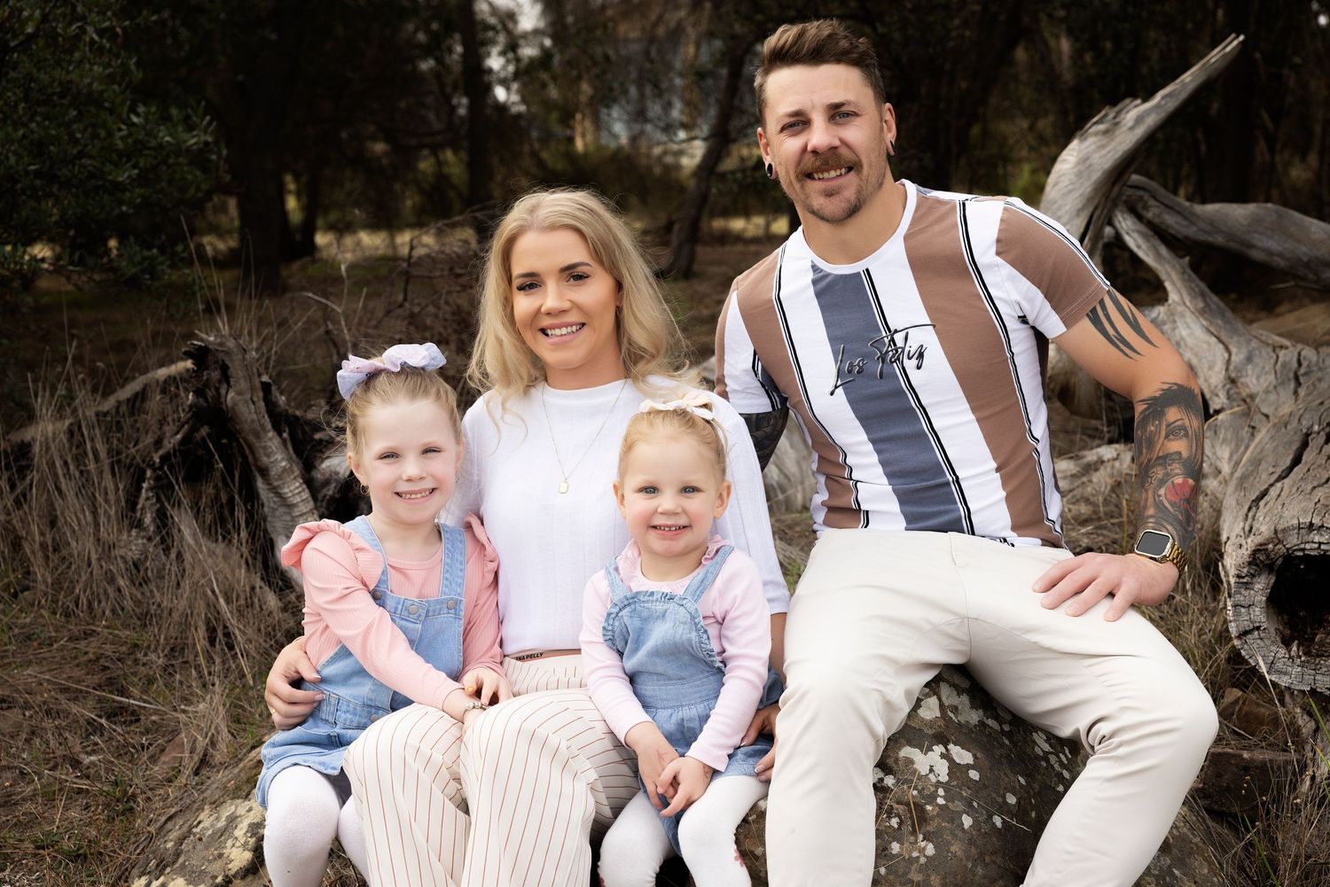 Happy family of four poses together on rustic driftwood in natural outdoor setting.