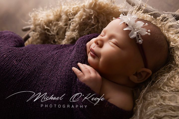 Newborn baby girl sleeping peacefully wrapped in purple, wearing a white flower headband, by Michael O'Keefe Photography.