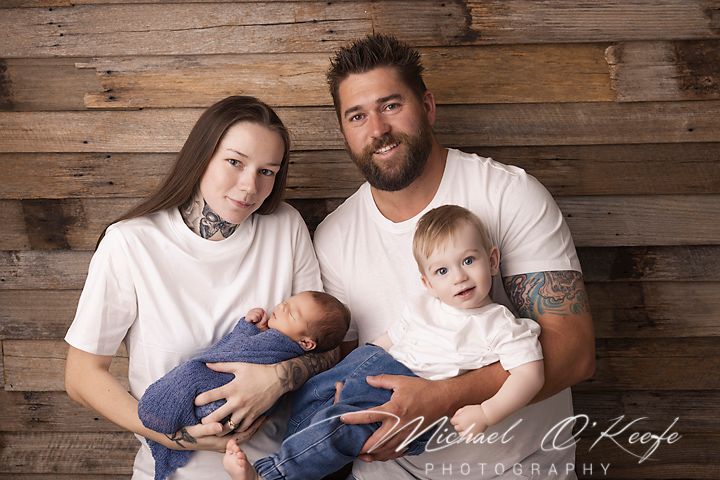 A family wearing white and blue sits together against a rustic wood plank wall for a portrait session.