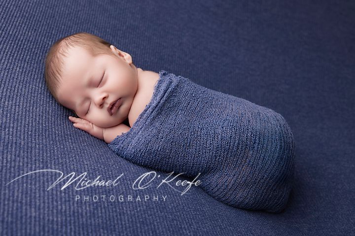 A sleeping newborn wrapped in a soft gray swaddle blanket rests peacefully on a navy blue textured backdrop.