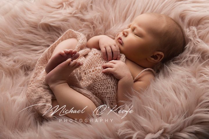Sleeping newborn baby rests peacefully on soft pink fur blanket while wearing delicate knit wrap during professional photoshoot.