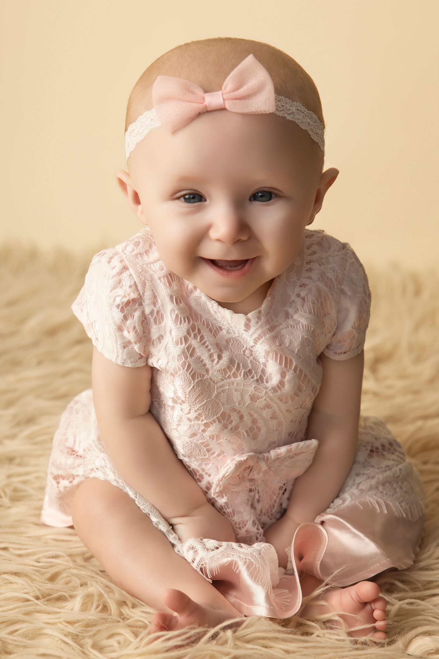 Smiling baby girl in pink lace dress and bow headband sitting on fluffy beige rug for portrait photo.