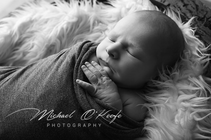 Black and white newborn photography by Michael O'Keefe showing a sleeping baby swaddled in soft wrap on fluffy rug.