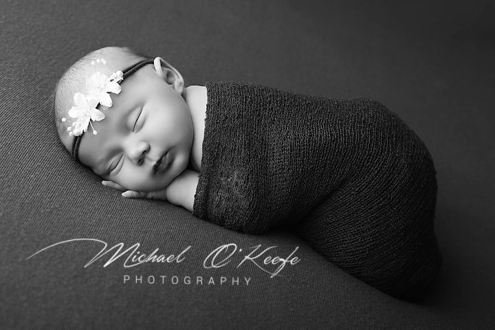 Black and white newborn portrait of sleeping baby swaddled in dark wrap with floral headband by Michael O'Keefe Photography.