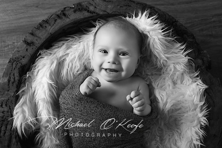 Black and white photo of a smiling baby lying in a fluffy bowl by Michael O'Keefe Photography.