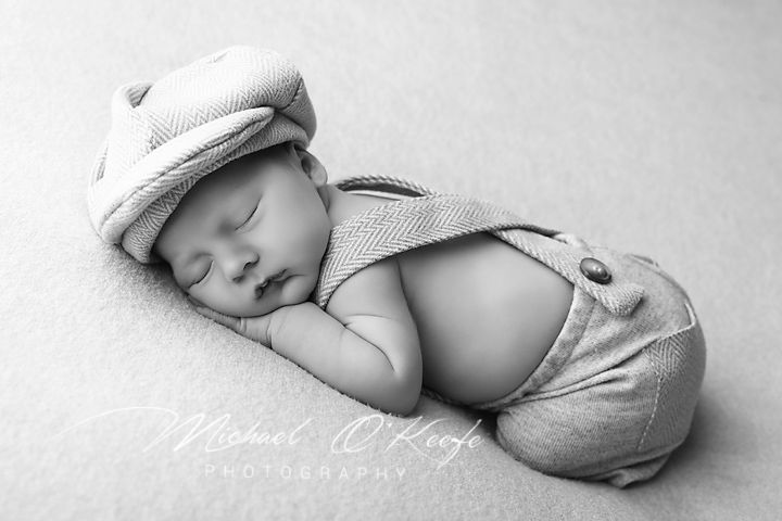 Black and white newborn photo of sleeping baby in knit cap and suspender outfit by Michael OKraft Photography.