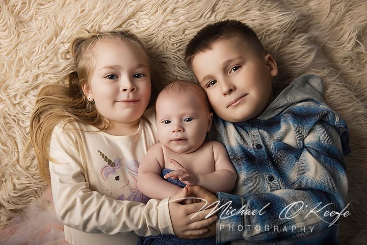Two siblings lovingly hold their newborn baby sibling on a soft fur backdrop in a professional family portrait.