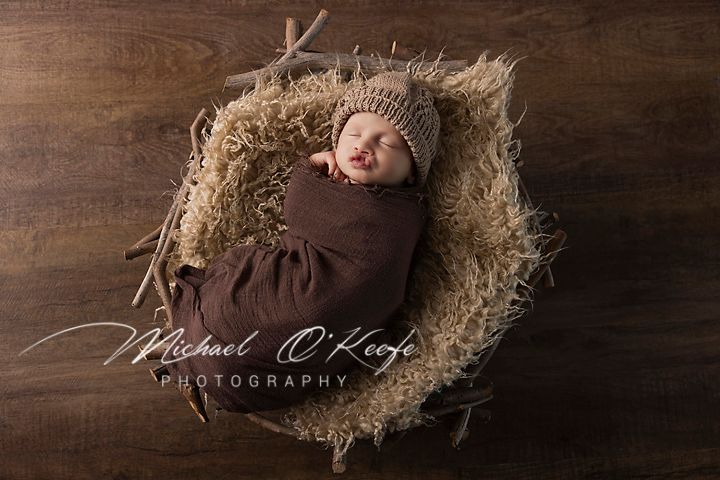 Newborn baby wrapped in brown blanket sleeping peacefully on wooden backdrop with straw nest arrangement.