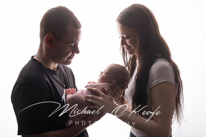 Young parents smiling and cradling their newborn baby in a professional white background studio portrait photo.