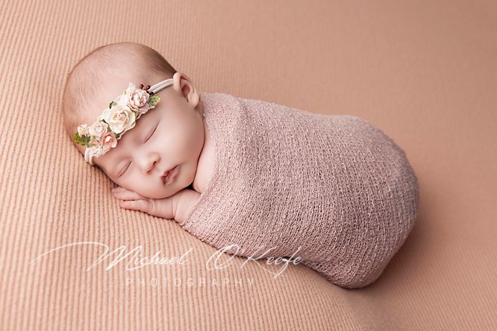 Peaceful newborn wrapped in pink swaddle wearing floral headband.