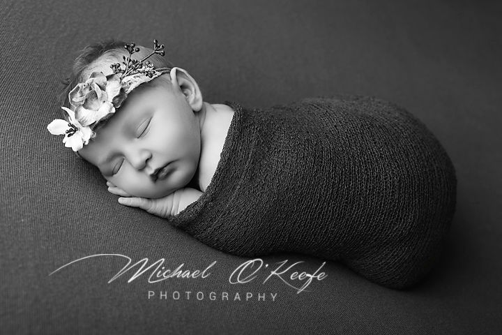 Black and white newborn photo of sleeping baby swaddled in dark wrap wearing floral headband by Michael O'Keefe Photography.