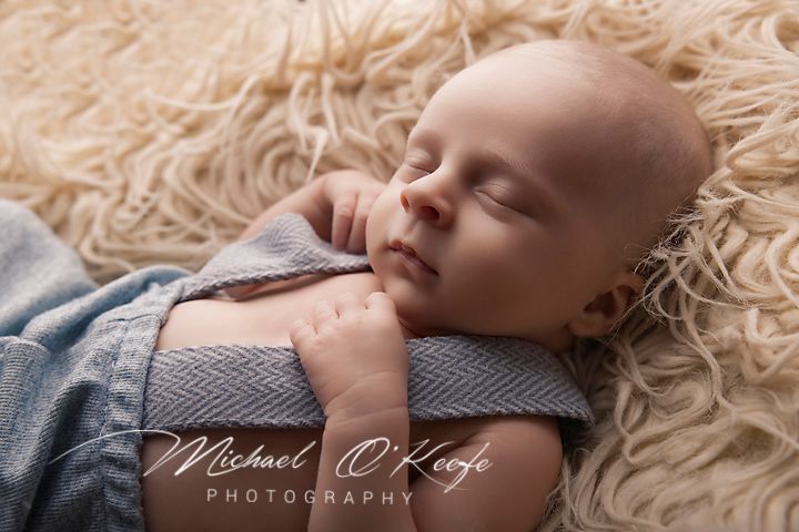 Peaceful newborn baby sleeping on a fluffy cream rug wrapped in a soft blue blanket, photographed by Michael OKeefo.