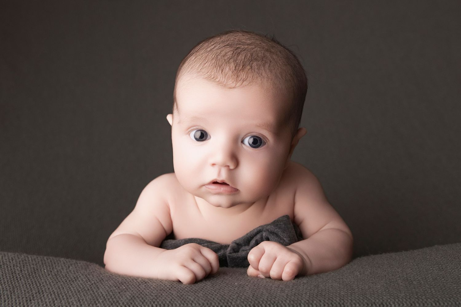 Adorable baby with blue eyes lying on tummy on a gray surface, looking curiously at the camera.