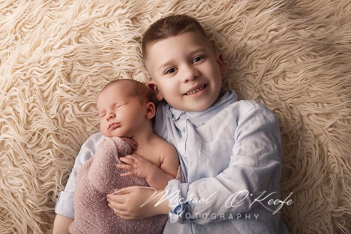 Older boy smiling while lying next to sleeping newborn baby wrapped in pink on a fluffy beige blanket.