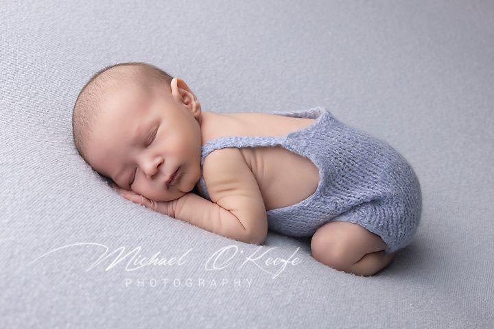 Sleeping newborn wearing blue knit romper rests peacefully on soft gray backdrop during professional photo session.