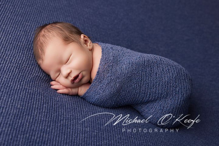 A sleeping newborn baby wrapped in a soft blue blanket rests peacefully on a navy blue textured surface.