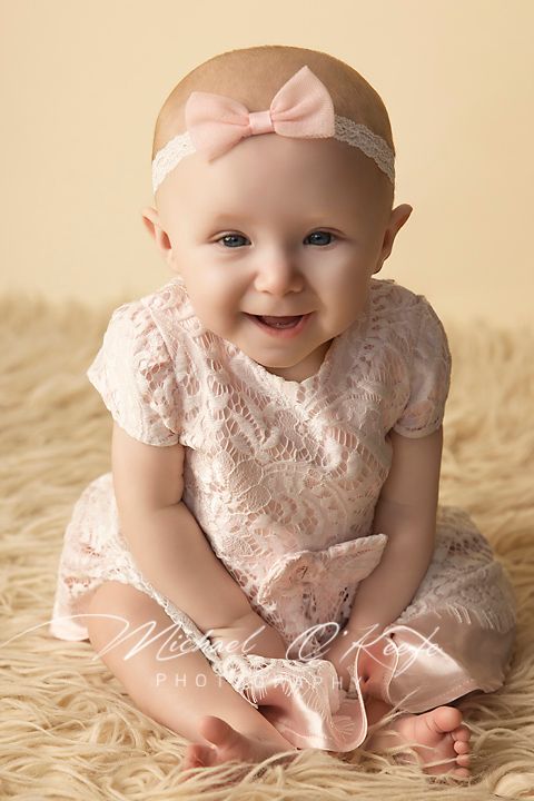Smiling baby girl in pink lace dress and bow headband sitting on fluffy rug for professional portrait photo.
