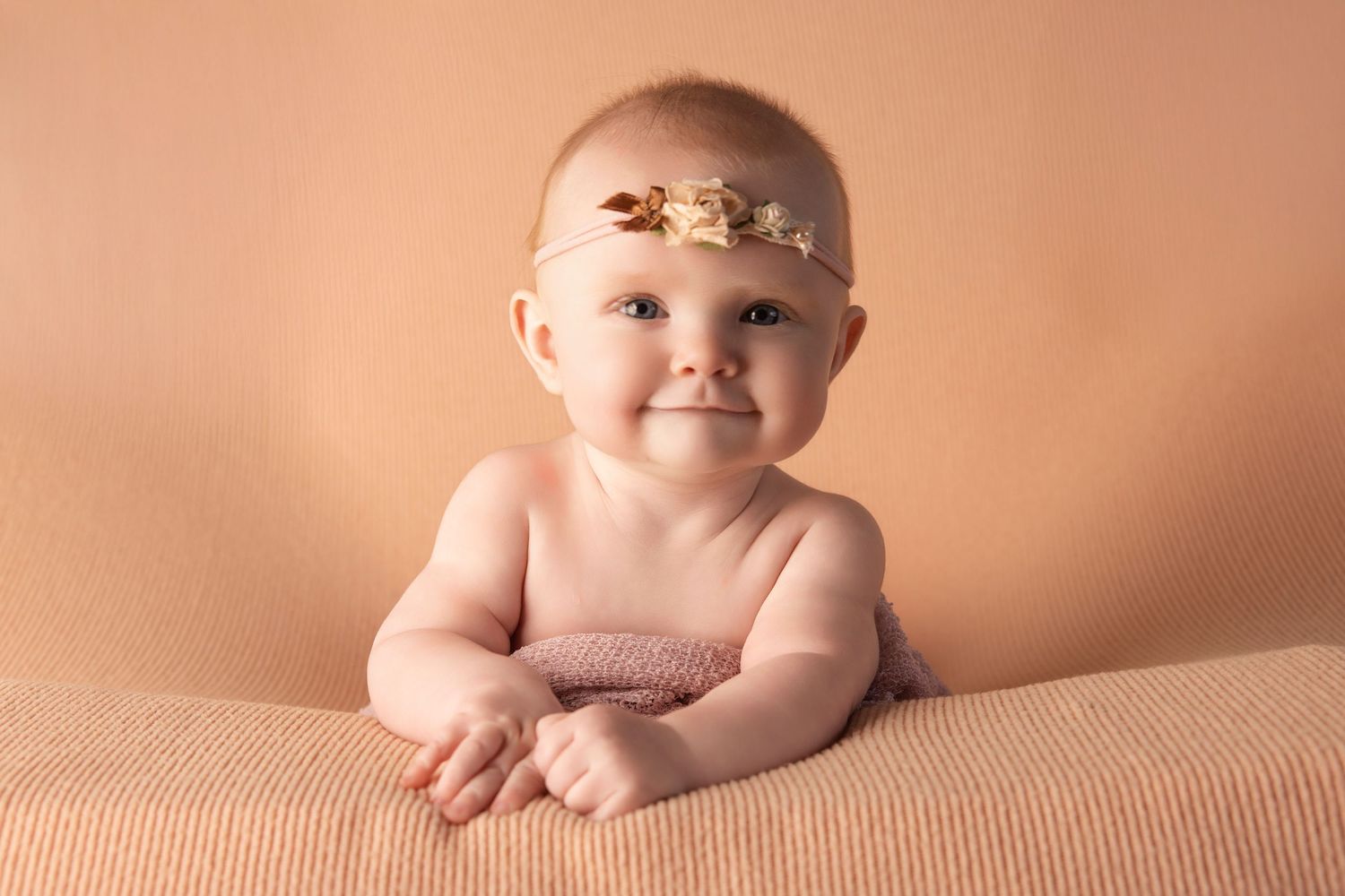 Baby wearing delicate floral headband sits on peach-colored backdrop.
