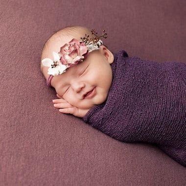 Sleeping newborn wrapped in purple swaddle with pink flower headband on mauve background.