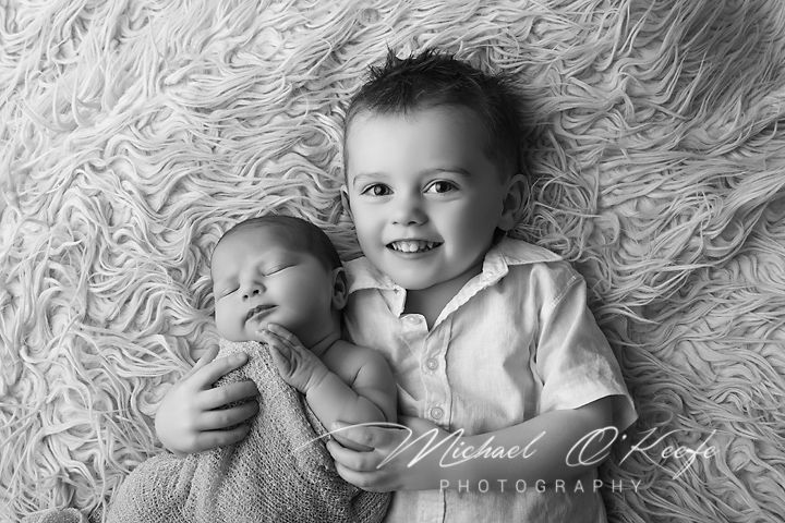 Black and white photo of a smiling toddler boy gently holding a sleeping newborn baby on a fluffy rug.