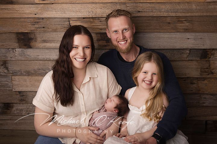 Family of four posing for a professional portrait, parents holding newborn baby with young daughter, rustic wood backdrop.