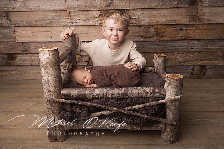Young boy smiling beside sleeping newborn in rustic log bed, professional newborn photography by Michael O'Keefe.