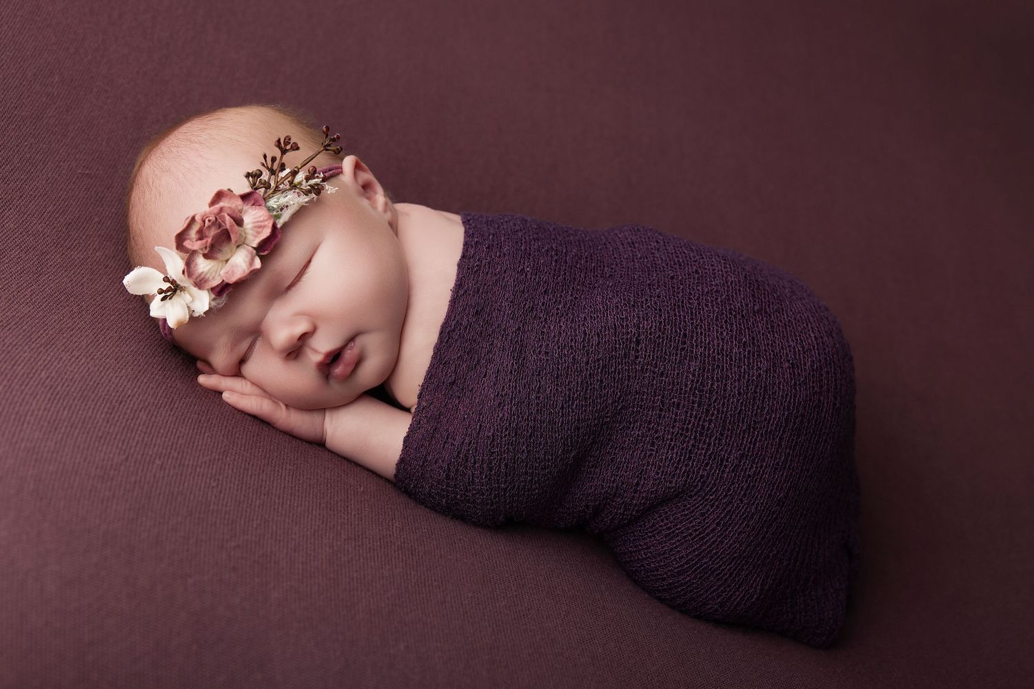 Sleeping newborn baby girl swaddled in purple wrap with floral headband on matching purple background.