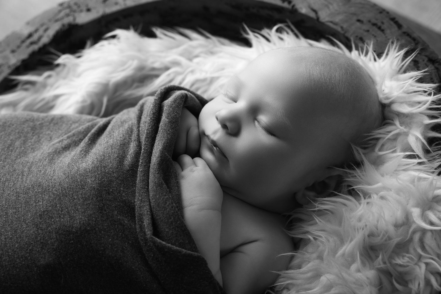 Peaceful newborn baby sleeping wrapped in soft blanket inside a rustic wooden bowl with fluffy fur lining.