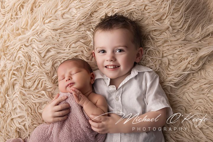 Smiling toddler boy gently holds sleeping newborn baby sibling on a soft cream fluffy rug in a studio photo.