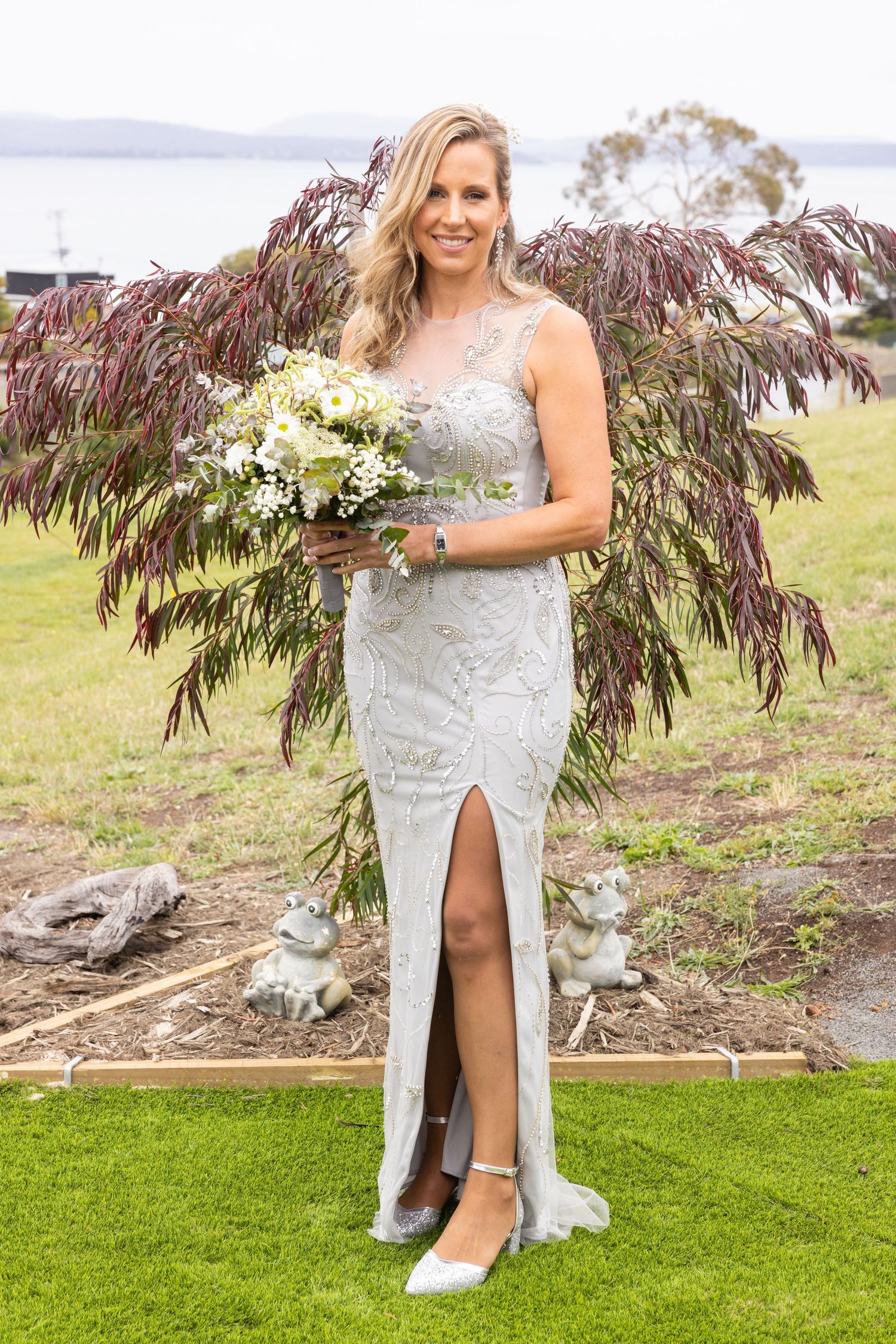 Bride in lace gown holds bouquet while standing on lawn with water and purple foliage backdrop.