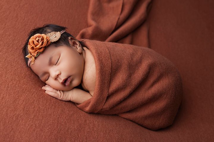 Sleeping newborn wrapped in rust-colored swaddle with matching floral headband.