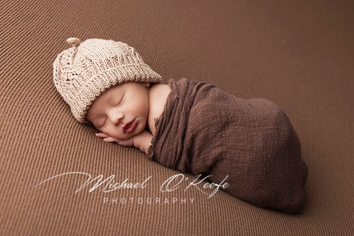 Newborn baby sleeping peacefully wrapped in brown swaddle blanket with knit beanie on brown backdrop.