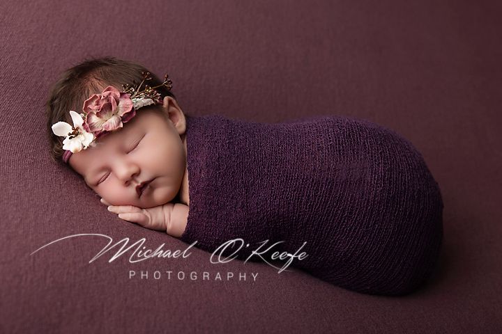 Newborn baby swaddled in purple wrap with floral headband, sleeping peacefully on purple background.