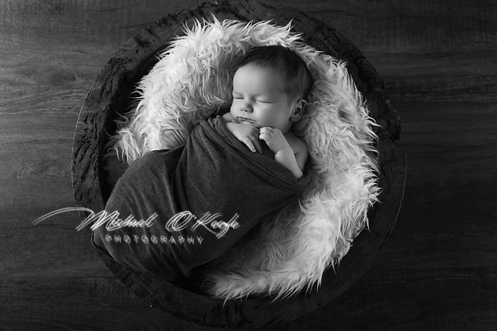 Black and white newborn photo of sleeping baby wrapped in gray swaddle nestled in fluffy white fur bowl prop.
