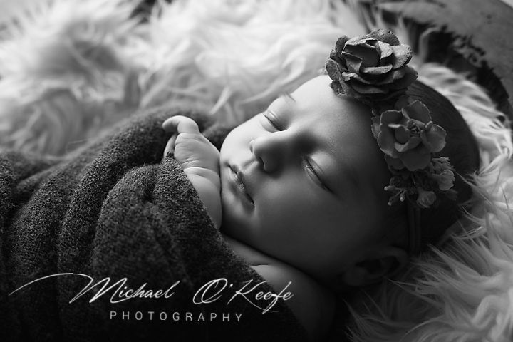 Black and white newborn photo of sleeping baby girl with floral headband, wrapped in soft blanket on fluffy white rug.