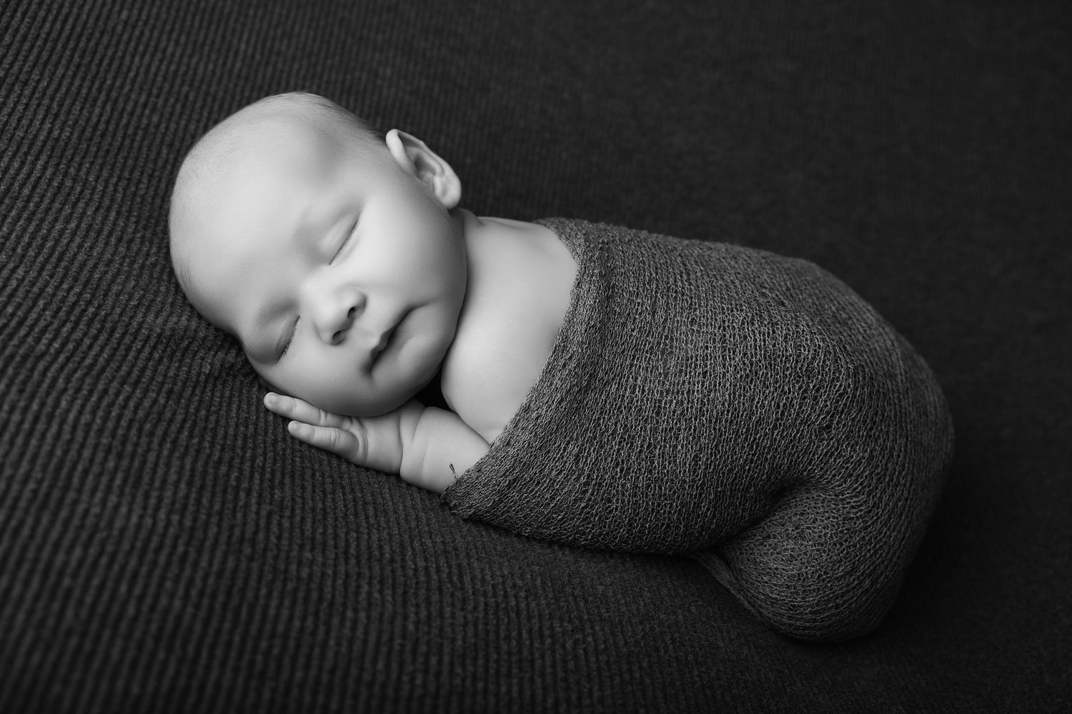 Black and white newborn photo of a sleeping baby swaddled in a knit wrap, posed on a dark textured backdrop.