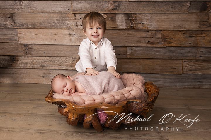 Newborn baby sleeping in wooden bowl with smiling toddler sibling, professional studio photography by Michael O'Keefe.