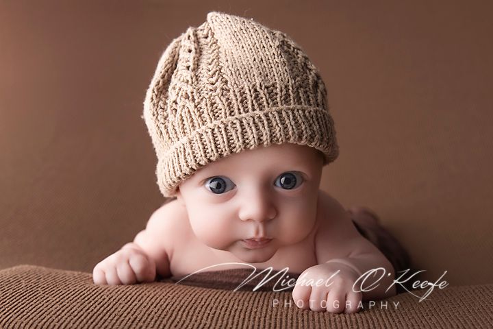Adorable baby wearing a knit beige hat lying on a brown blanket, looking up with big blue eyes.