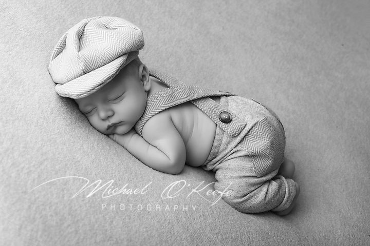 Black and white newborn photo of sleeping baby wearing knit cap and suspender outfit by Michael O'Keefe Photography.