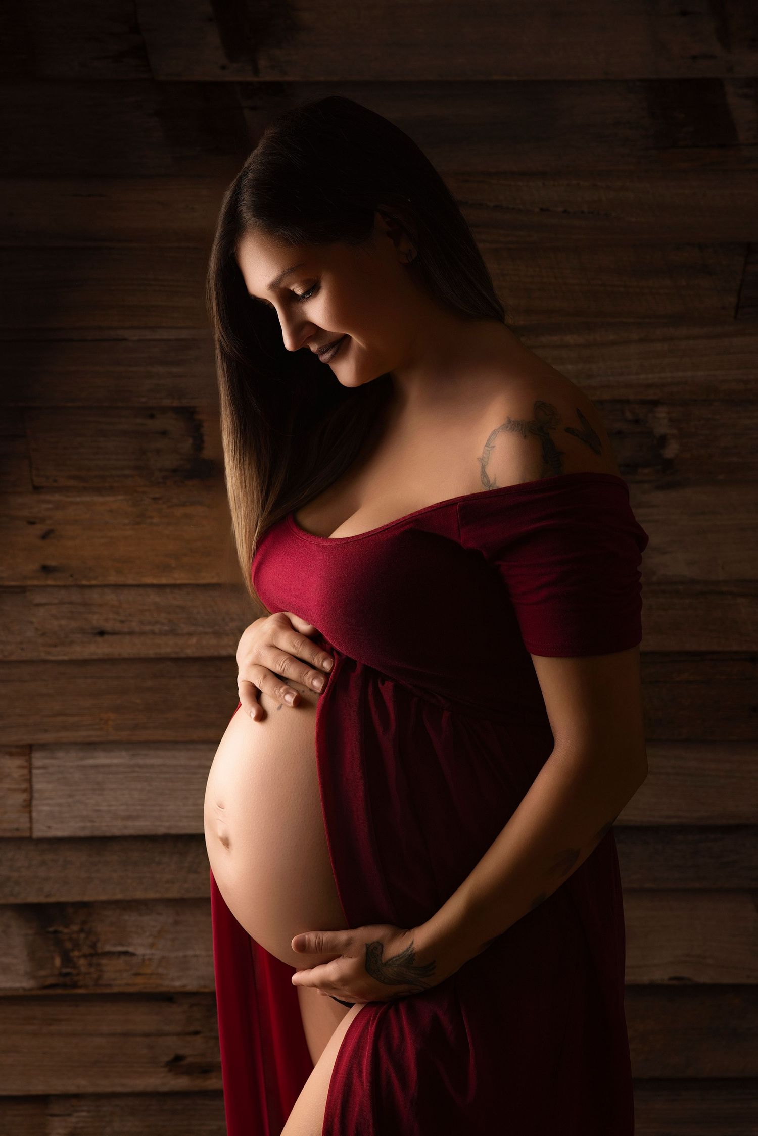 Maternity portrait features woman in flowing red dress against rustic wooden wall background.
