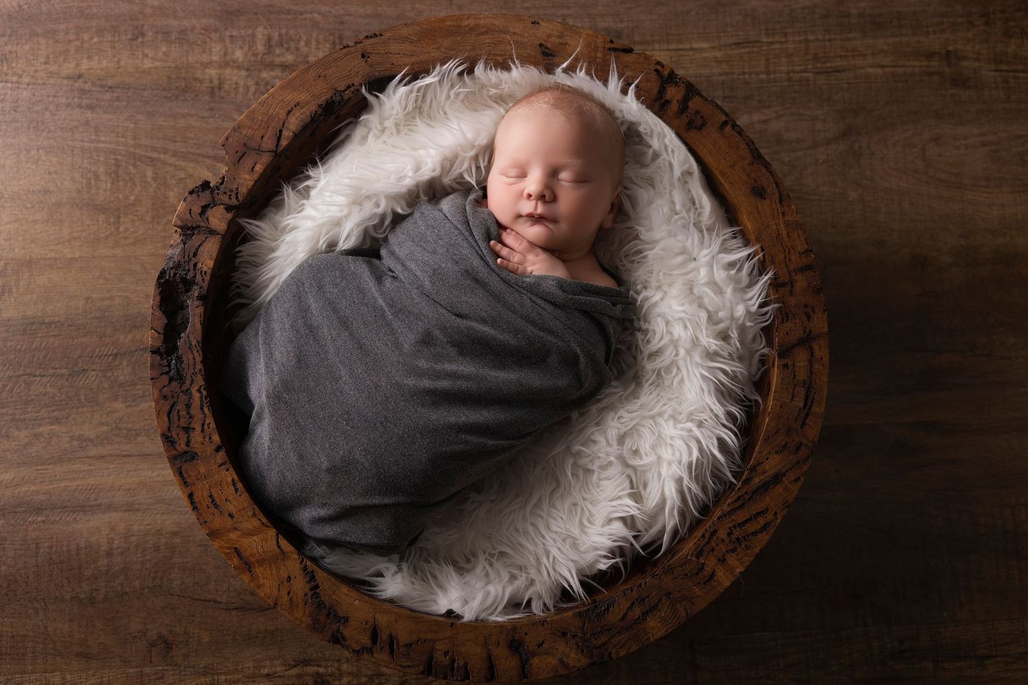 Sleeping newborn swaddled in gray wrap resting in wooden bowl with white fur.