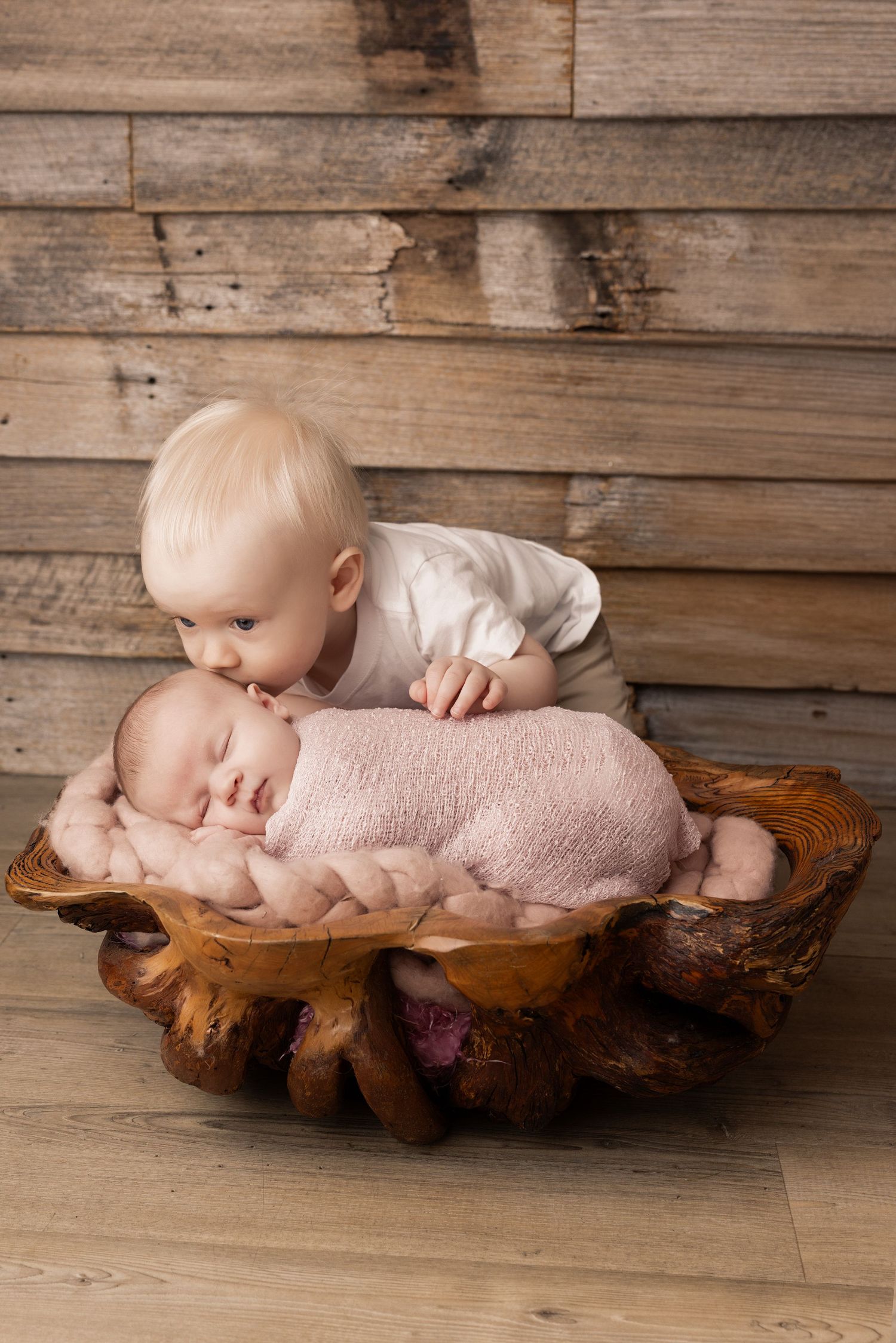 Toddler looking over sleeping newborn on wooden platform with rustic background.