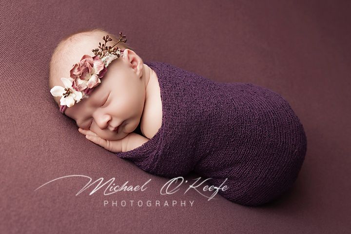 A newborn baby sleeps peacefully on a purple blanket in a professional studio photography setting.