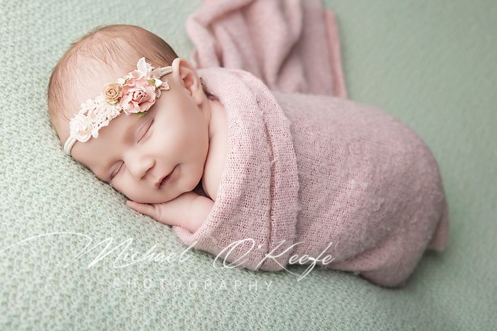 Newborn baby girl swaddled in pink wrap with floral headband sleeping peacefully on mint green background.